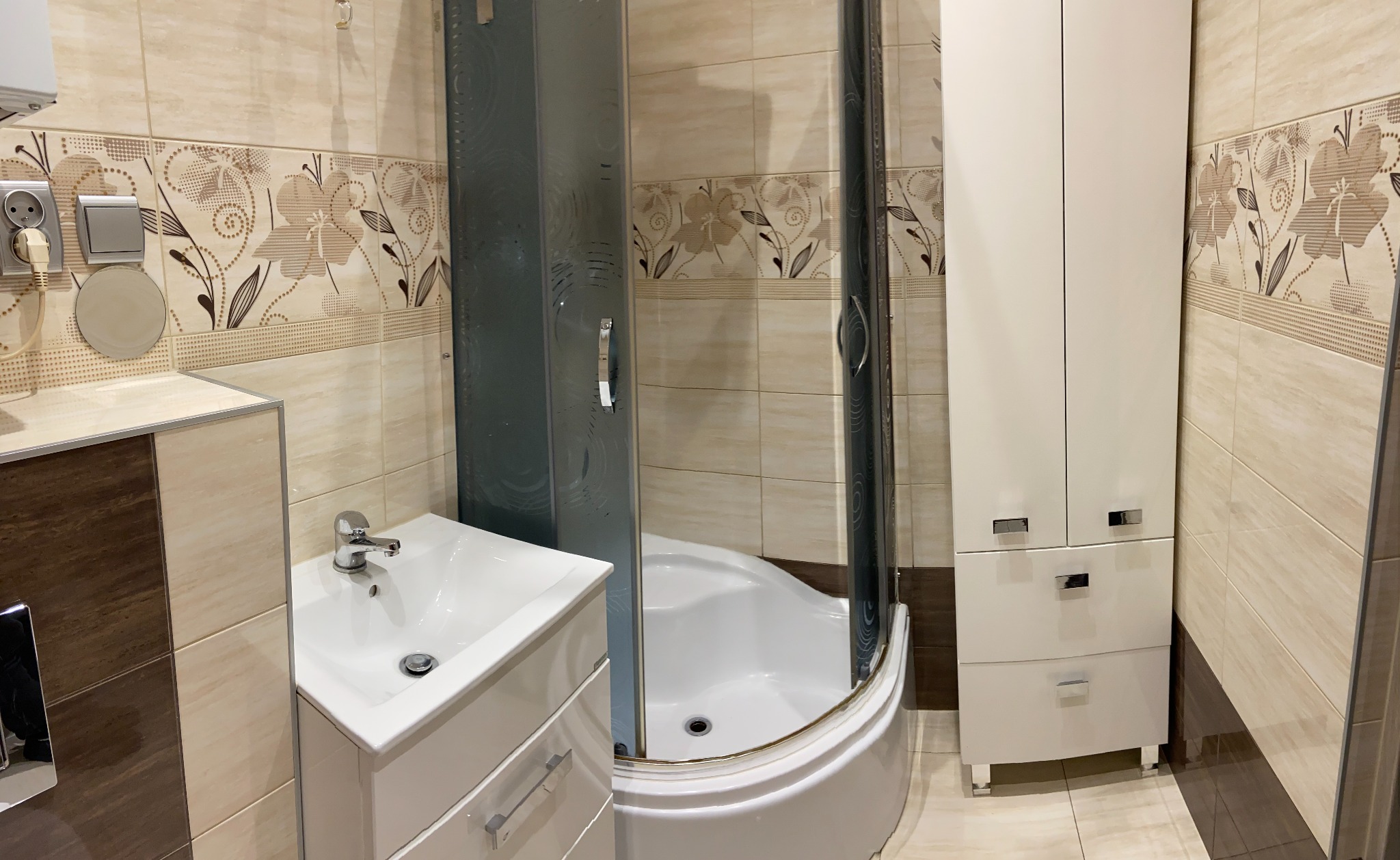 Wykończona łazienka z beżowymi płytkami w kwiaty, białą kabiną prysznicową z szarymi drzwiami, umywalką z szafką i wysoką szafką z lustrzanymi uchwytami.