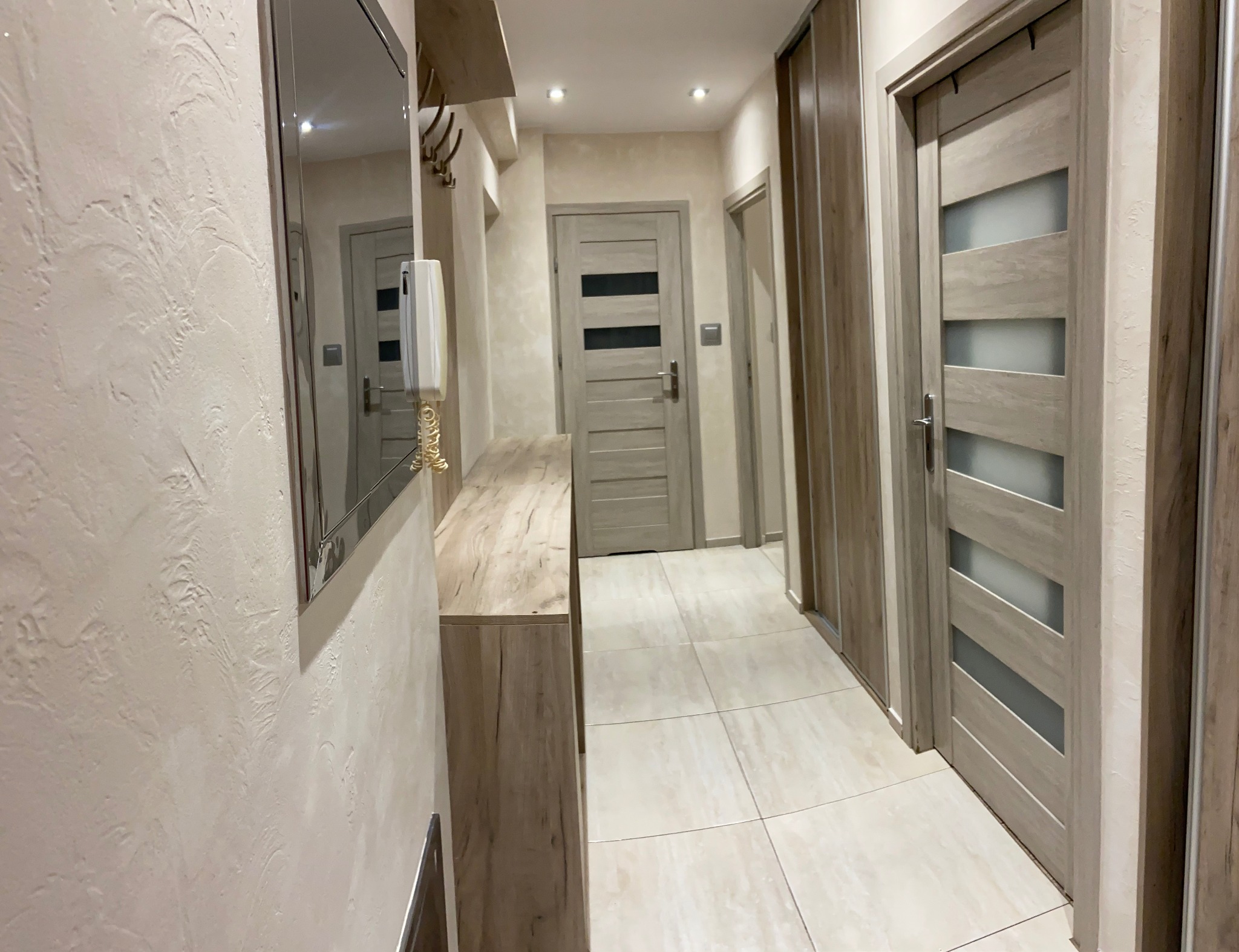 Jasne wnętrze przedpokoju z beżowymi płytkami podłogowymi, teksturowanymi ścianami, lustrem, drewnianą konsolą, wieszakiem na ubrania i trzema drzwiami w odcieniach szarości, z których jedne mają...