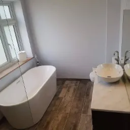 Nowoczesna łazienka z wolnostojącą wanną przy oknie, ceramiczną umywalką nablatową i podłogą wyłożoną panelami imitującymi drewno.