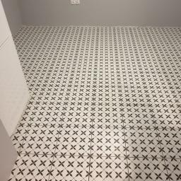 Usługi Budowlane Kubasik - Ułożona podłoga z białych płytek w czarny wzór kwiatowy, widoczna krawędź białej lodówki i szara ściana w tle.