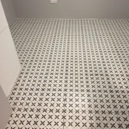 Ułożona podłoga z białych płytek w czarny wzór kwiatowy, widoczna krawędź białej lodówki i szara ściana w tle.