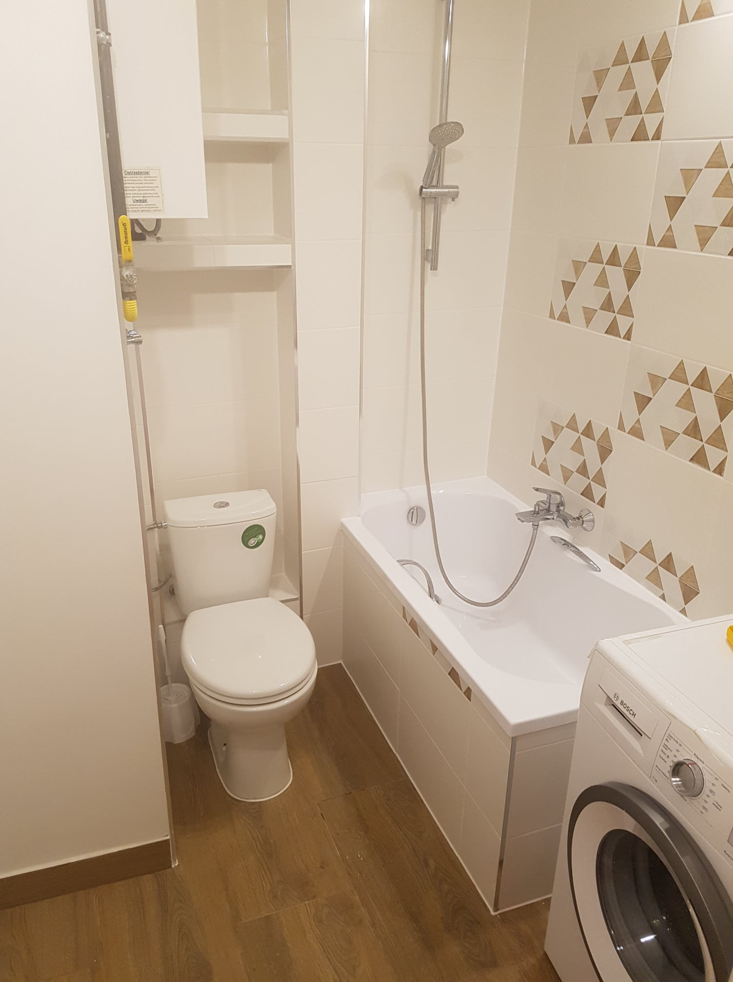 Jasna łazienka z toaletą, wanną obudowaną kafelkami z dekoracyjnym brązowym wzorem trójkątów, pralką Bosch i drewnopodobną podłogą, oświetlona ciepłym światłem.