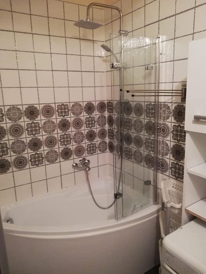 Narożna wanna z chromowaną armaturą i szklanym parawanem w łazience wykończonej białymi płytkami z dekoracyjnym, ciemnym wzorem.