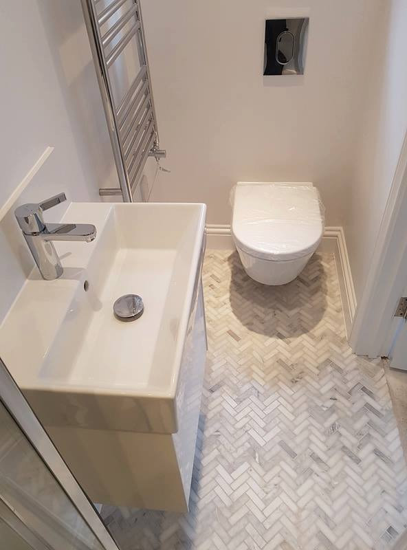 Nowoczesna, mała łazienka z białą umywalką na szafce, podwieszaną toaletą w folii ochronnej i grzejnikiem drabinkowym, wykończona marmurową mozaiką ułożoną w jodełkę.