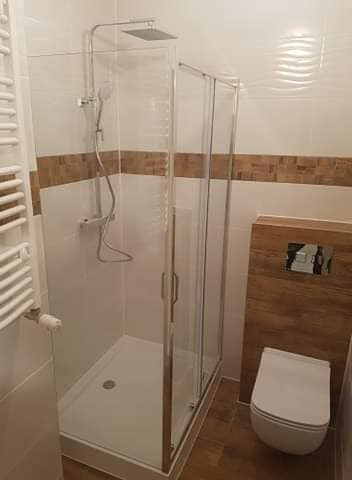 Nowoczesna kabina prysznicowa z deszczownicą i przesuwanymi drzwiami, widoczna obok podwieszana toaleta z drewnianą obudową w łazience wykończonej białymi płytkami z brązowym pasem dekoracyjnym.