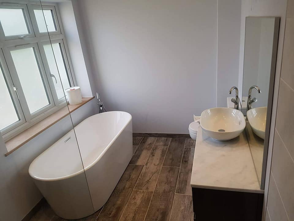 Nowoczesna łazienka z wolnostojącą wanną przy oknie, ceramiczną umywalką nablatową i podłogą wyłożoną panelami imitującymi drewno.