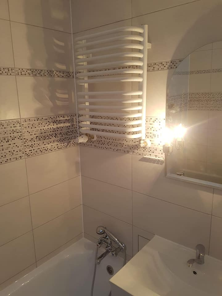 Biały grzejnik drabinkowy zawieszony na ścianie wyłożonej jasnymi płytkami z dekoracyjnym pasem mozaiki w łazience z wanną i umywalką.