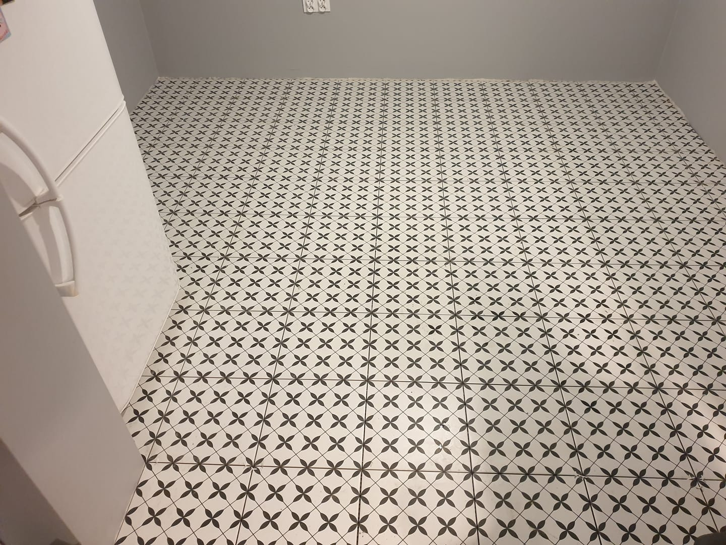 Ułożona podłoga z białych płytek w czarny wzór kwiatowy, widoczna krawędź białej lodówki i szara ściana w tle.