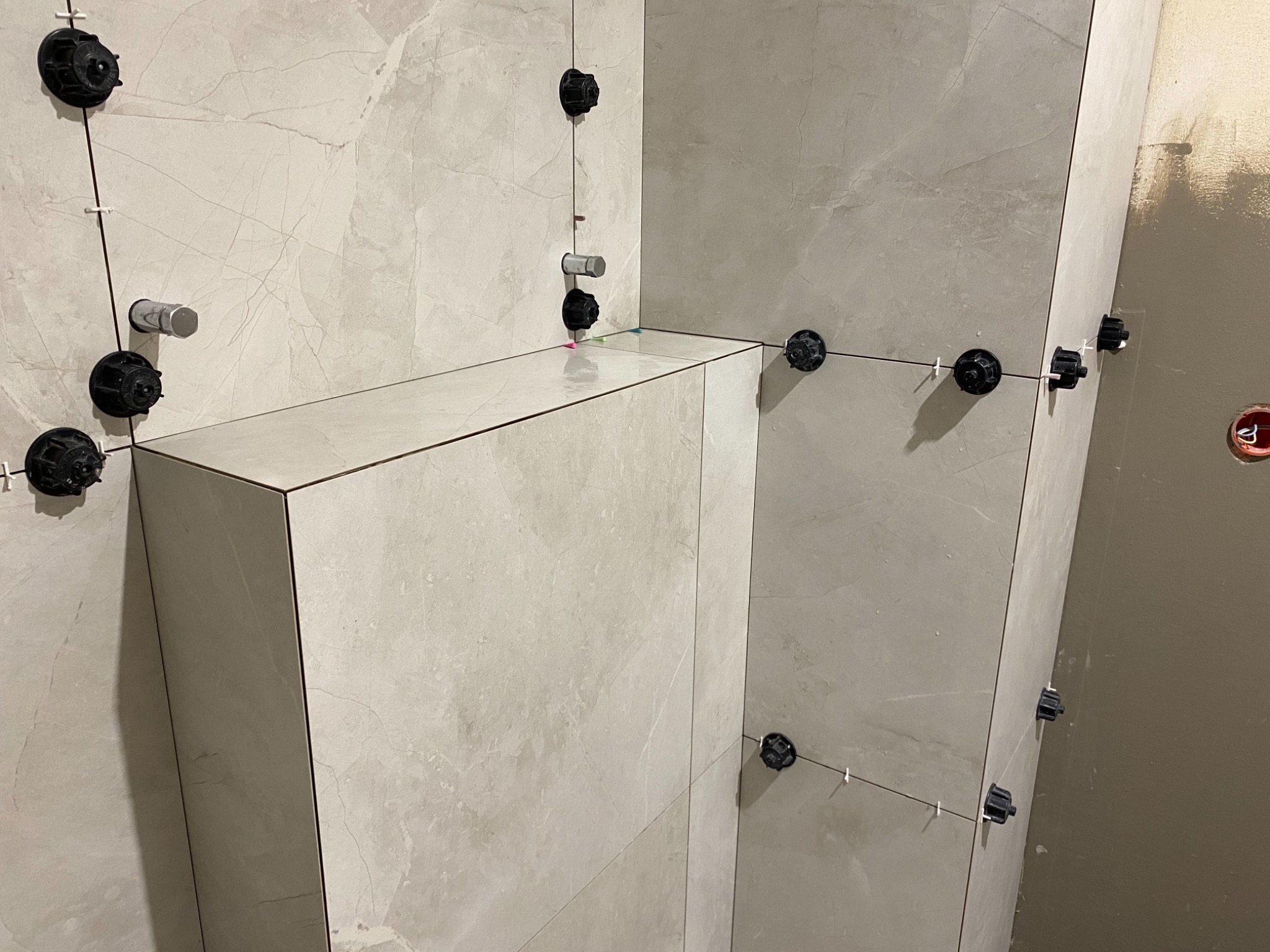 Montaż beżowych płytek wielkoformatowych z użyciem systemu poziomowania płytek, widoczne elementy instalacji wodnej i ściana przygotowana do malowania.