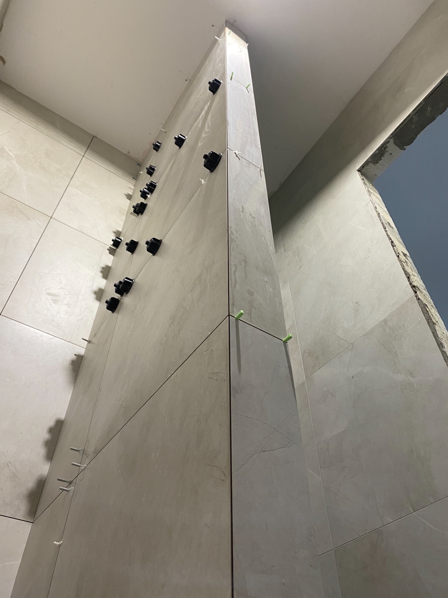 Ściana w trakcie układania płytek ceramicznych w odcieniach beżu i szarości, z użyciem czarnych klipsów poziomujących i zielonych krzyżyków dystansowych, widoczne surowe wykończenie otworu drzwiowego.