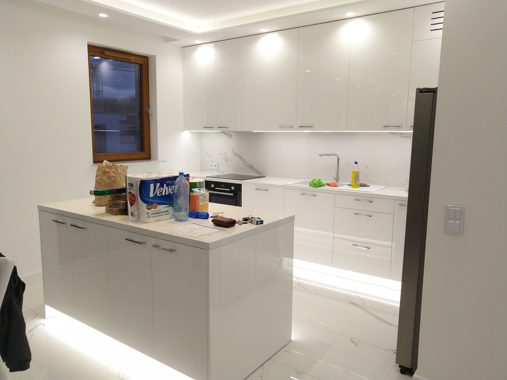 Nowoczesna, biała kuchnia z wyspą i marmurowym blatem, oświetlona listwami LED pod szafkami i na wyspie, widok na okno z widokiem na budowę.