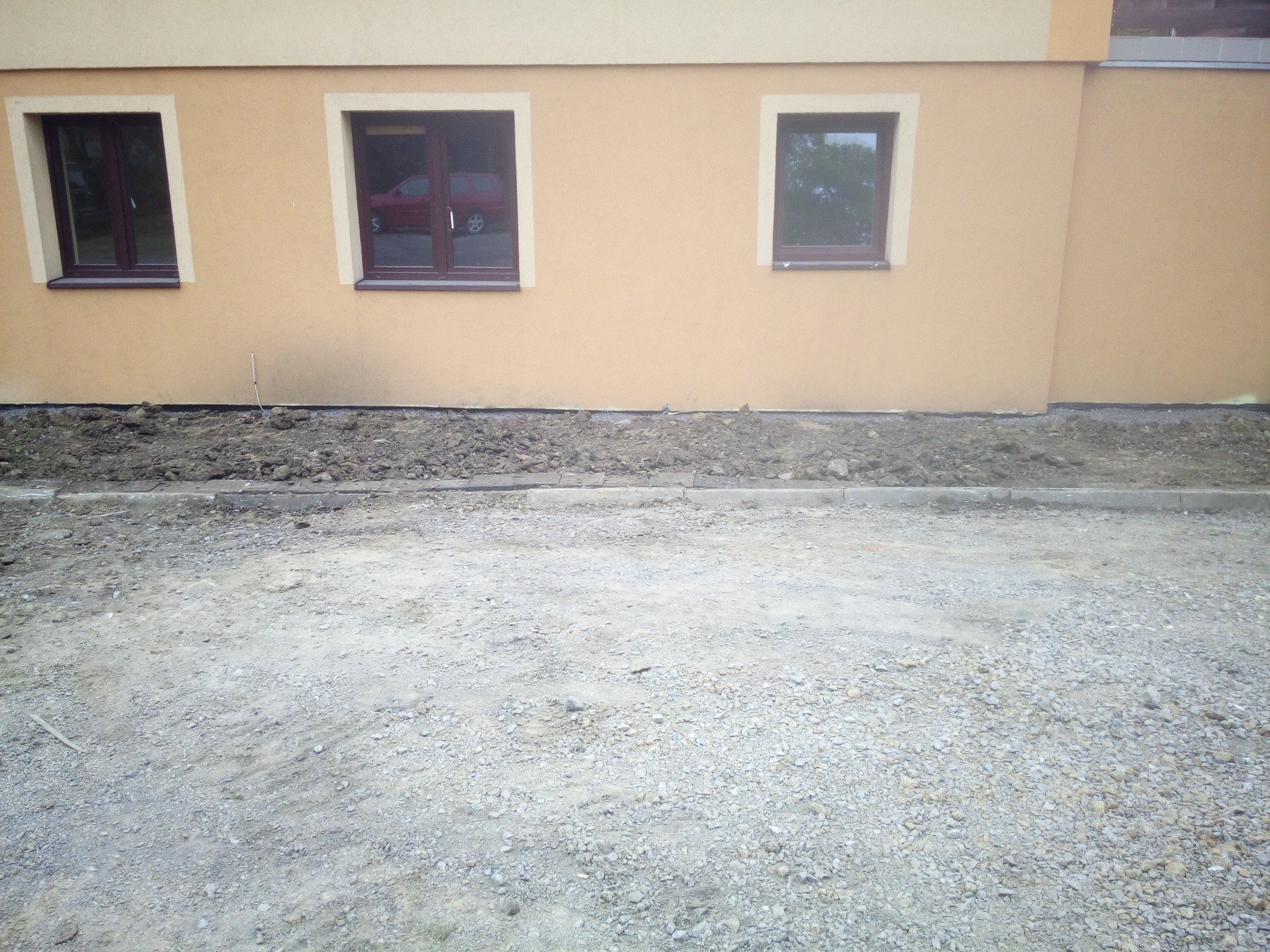 Przygotowany teren wokół budynku z trzema oknami; widoczna wykopana ziemia przy fundamencie i gruz na podłożu.