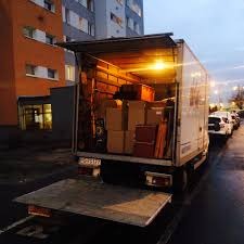 Otwarty tył ciężarówki wypełniony kartonami i meblami podczas wieczornej przeprowadzki, widoczny numer rejestracyjny pojazdu, budynek mieszkalny w tle.