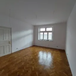 Pusty pokój po świeżym pomalowaniu ścian na biało, widoczna drewniana podłoga w jodełkę, białe okno i białe drzwi. Z sufitu zwisają przewody.