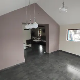 Wnętrze po remoncie: ściany w odcieniach beżu i lila, ciemna podłoga z płytek, widoczne gniazdko elektryczne, okno z widokiem na zewnątrz, w tle salon z meblami.