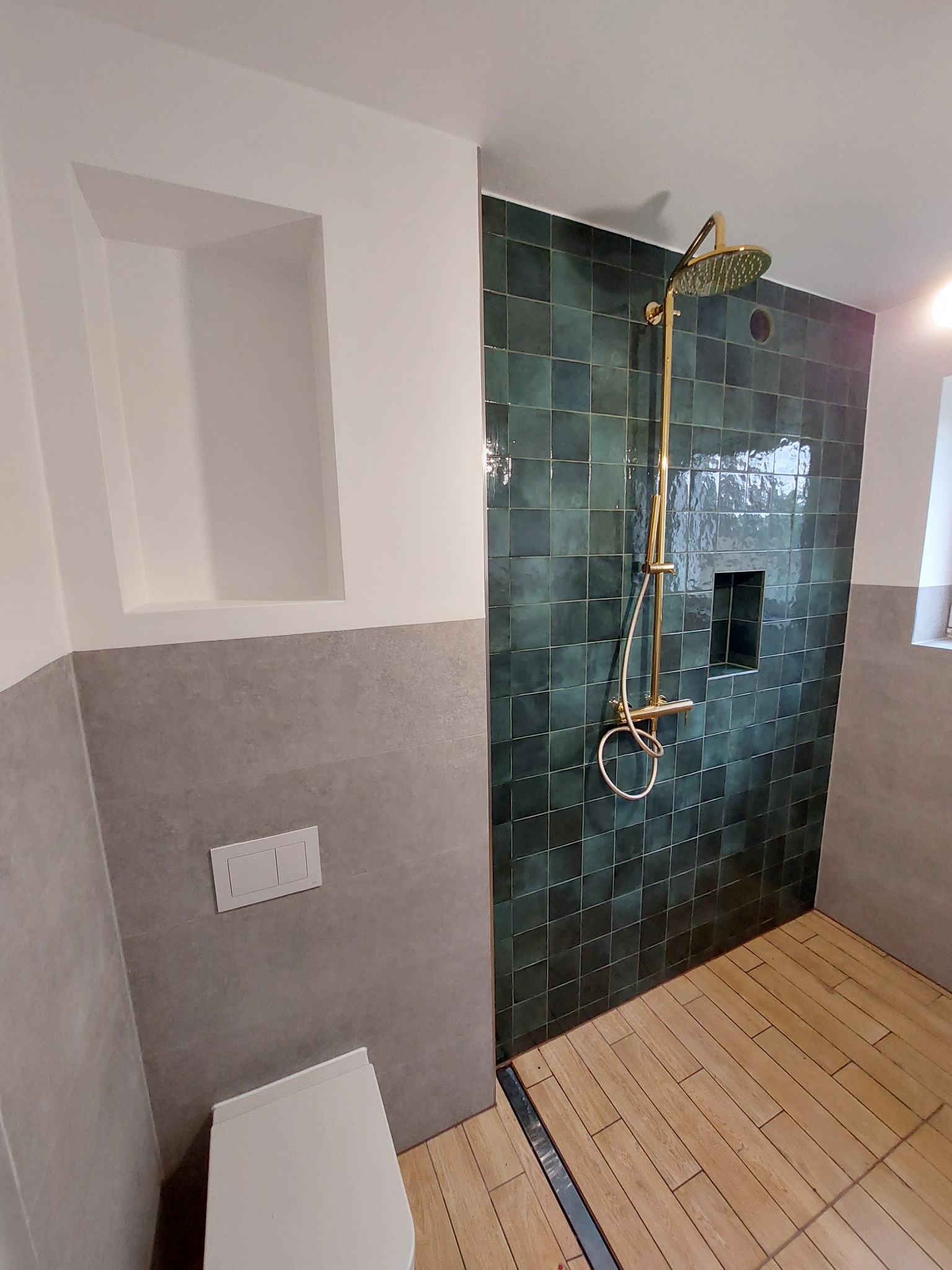 Nowoczesna łazienka z kabiną prysznicową wyłożoną ciemnozielonymi, kwadratowymi płytkami, złotą armaturą i drewnopodobną podłogą. W ścianie wnęka z półkami.