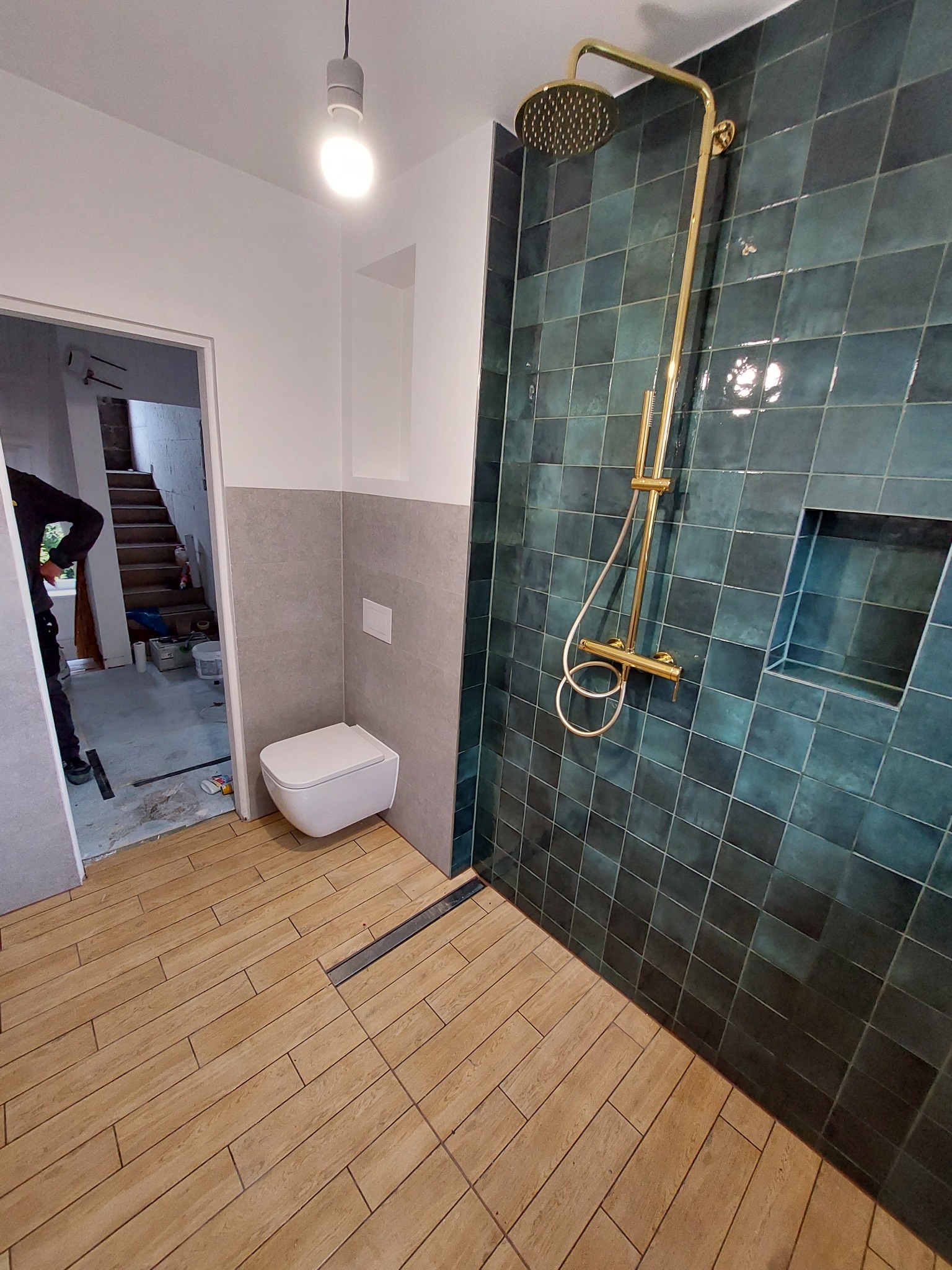 Nowoczesna łazienka w trakcie wykończenia: turkusowe płytki ścienne, złota armatura prysznicowa, drewnopodobne płytki podłogowe, wisząca toaleta i widoczne prace wykończeniowe w przejściu...