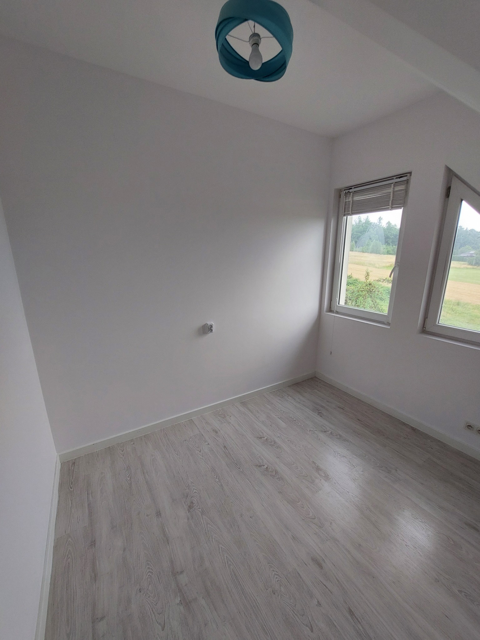 Pusty pokój po remoncie z białymi ścianami, panelami podłogowymi w odcieniach szarości i błękitną lampą sufitową. Widoczne dwa okna z widokiem na zieleń.