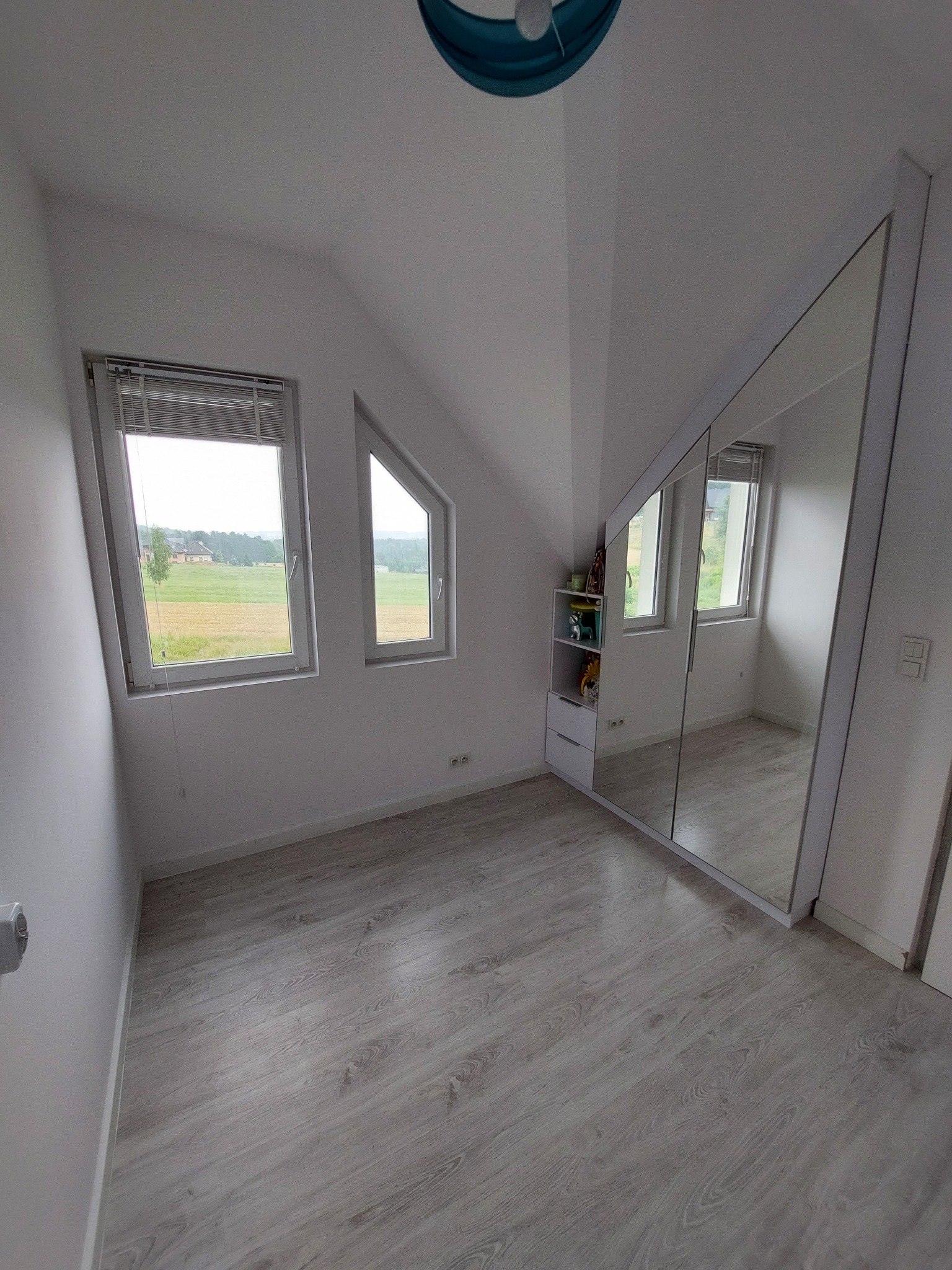 Jasne, puste poddasze po remoncie z drewnopodobną podłogą, skosami, szafą z lustrzanymi drzwiami i widokiem na pola za oknem.