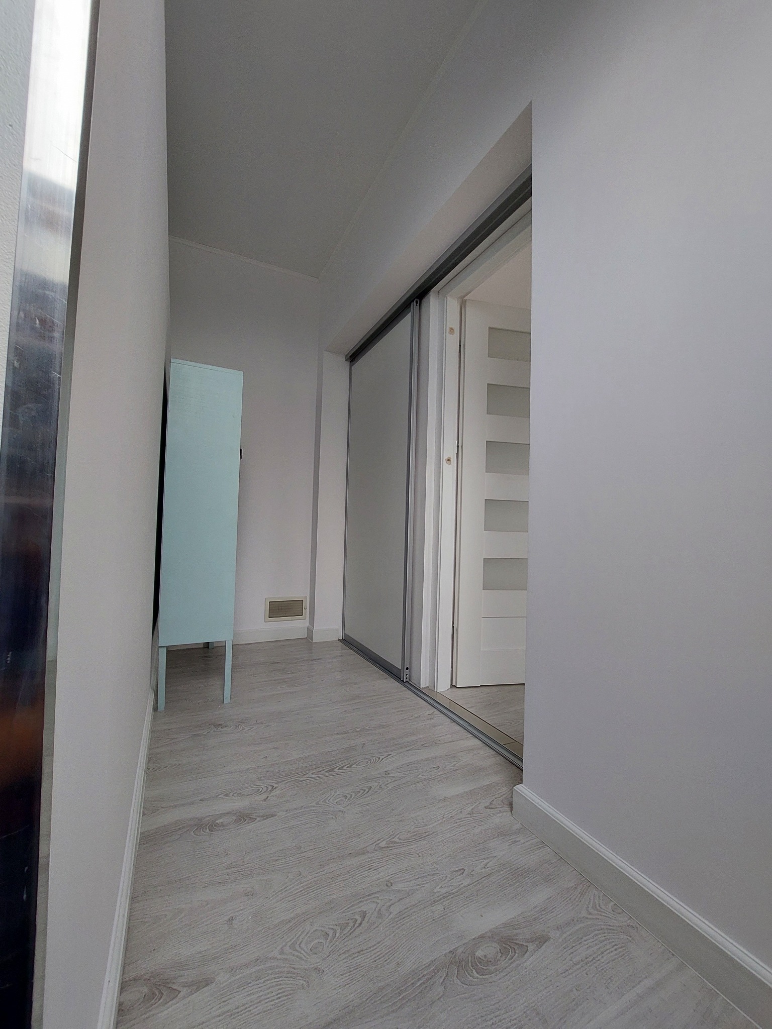 Korytarz z szarą podłogą imitującą drewno, jasnymi szarymi ścianami, przesuwnymi drzwiami z metalową ramą i niebieską szafką przy ścianie.