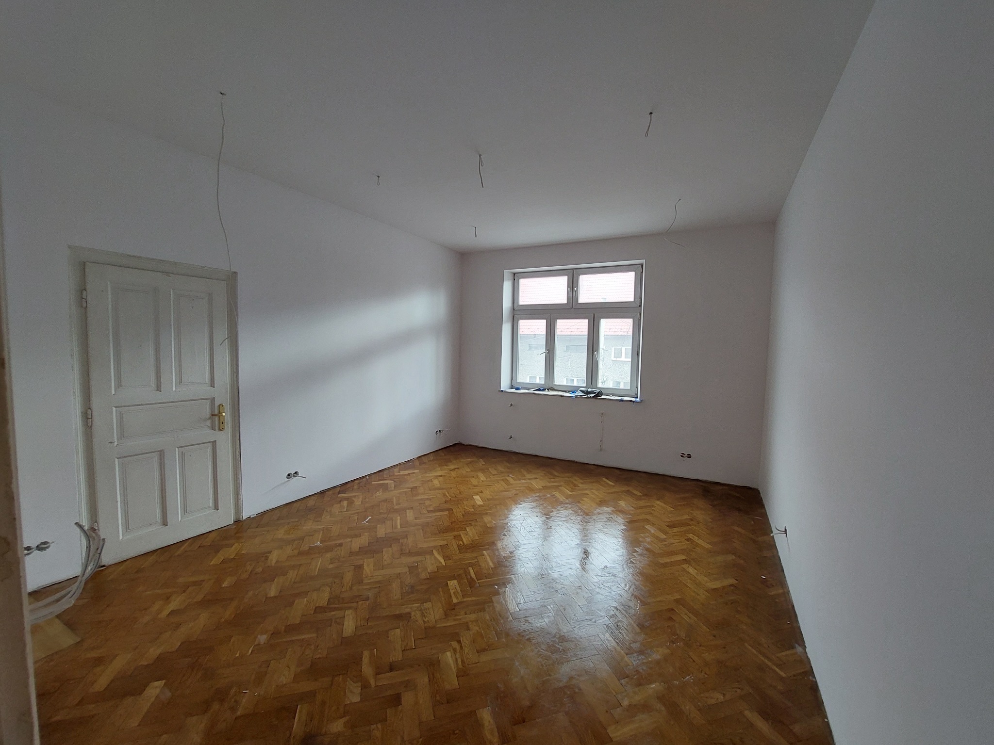 Pusty pokój po świeżym pomalowaniu ścian na biało, widoczna drewniana podłoga w jodełkę, białe okno i białe drzwi. Z sufitu zwisają przewody.