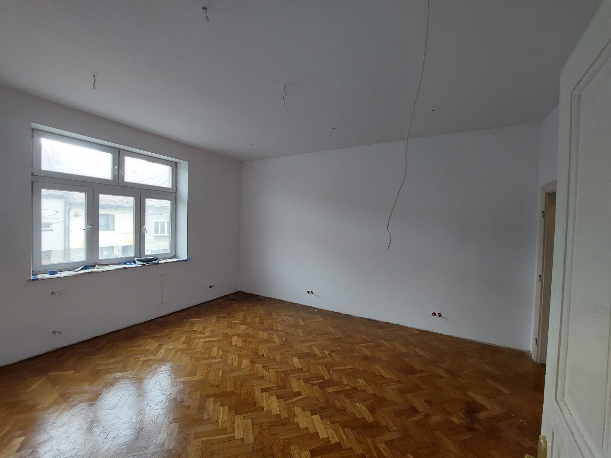 Pusty pokój po remoncie z parkietem w jodełkę, świeżo pomalowane ściany na biało, widoczne wystające przewody elektryczne i otwory po gniazdkach, okno z widokiem na budynki.