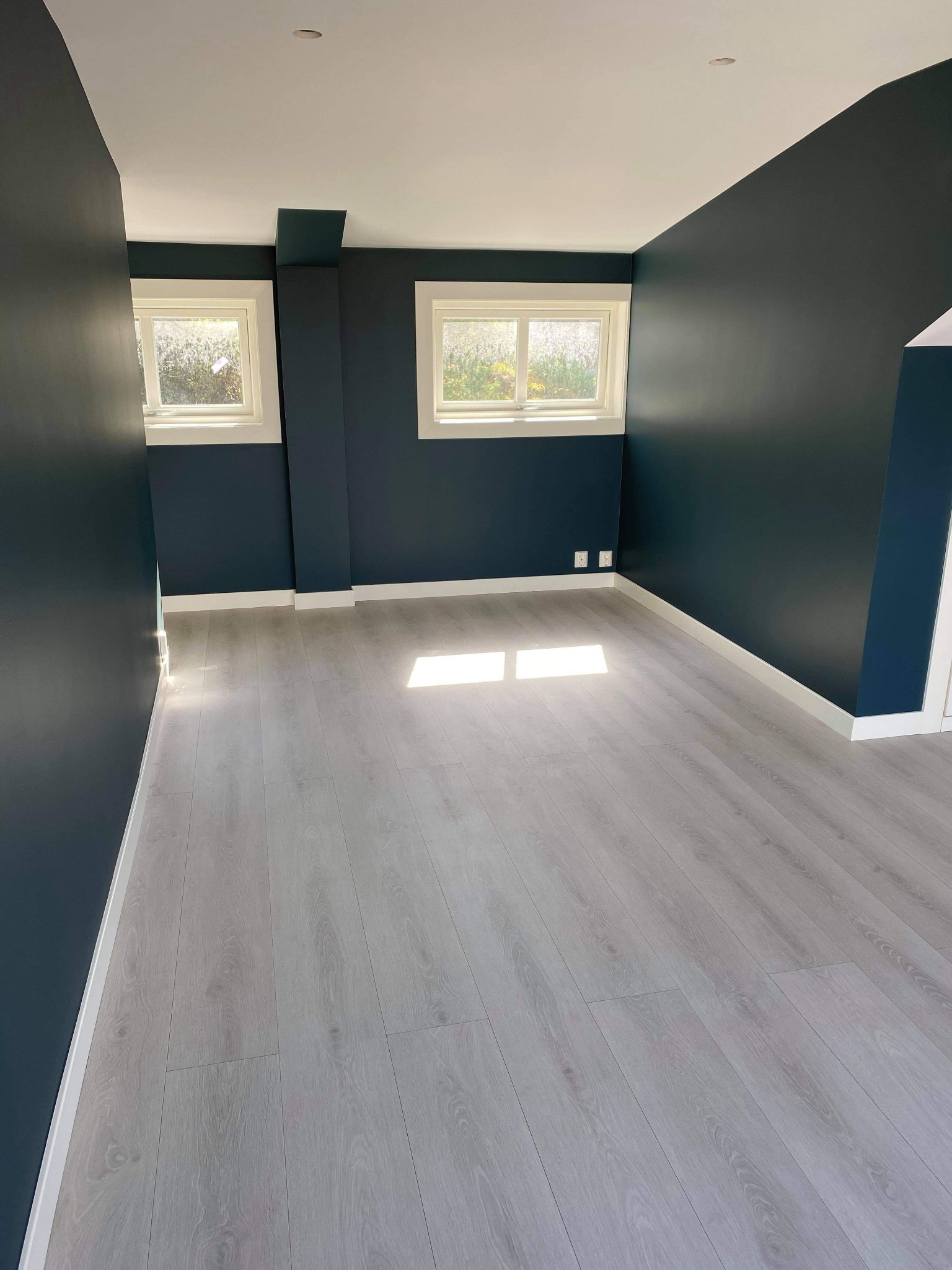 Pusty pokój po remoncie z pomalowanymi na ciemny granat ścianami, białym sufitem, dwiema oknami z białymi ramami i jasnoszarą podłogą z paneli.
