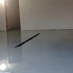 Świeżo wylana, szara posadzka z odpływem liniowym w surowym pomieszczeniu z oknem i instalacjami na ścianie.