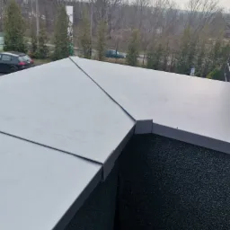 Szary, płaski dach z blachy na budynku z czarnym, chropowatym wykończeniem ściany, widok z góry w pochmurny dzień.