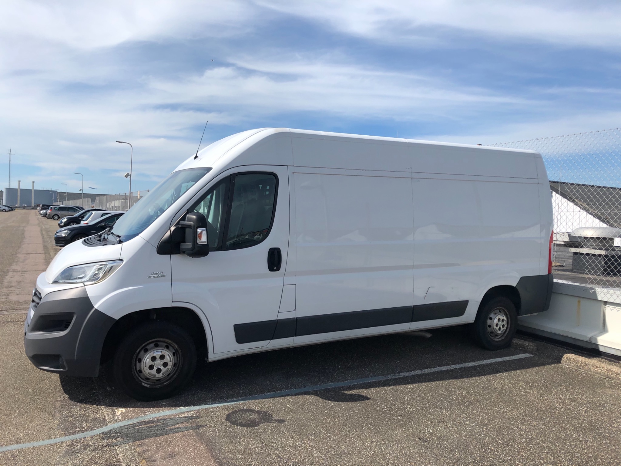 Biały furgon dostawczy Fiat Ducato zaparkowany na parkingu z widocznymi innymi samochodami i budynkami w tle, sfotografowany w słoneczny dzień.