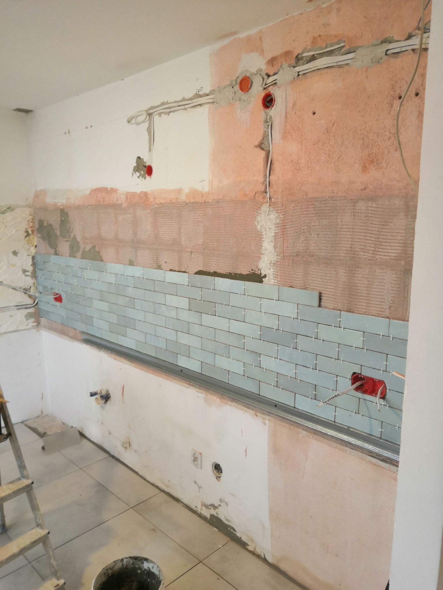 Montaż jasnoniebieskich płytek typu metro w kuchni, widoczne krzyżyki dystansowe, odsłonięte przewody elektryczne i fragment drabiny.