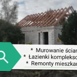 Budowa domu jednorodzinnego w stanie surowym otwartym: ściany z bloczków i drewniana konstrukcja dachu na tle zieleni.