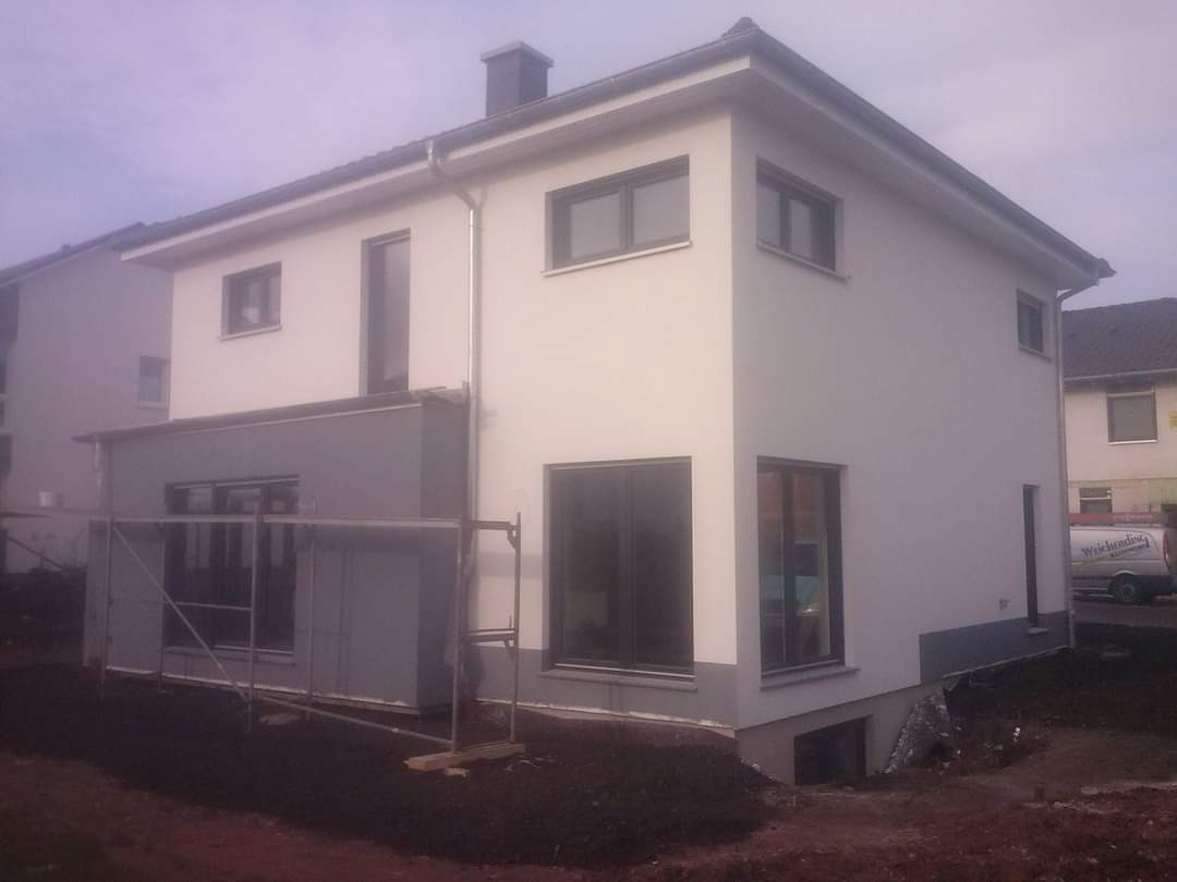 Dwupiętrowy dom jednorodzinny z nową, jasną elewacją i szarym cokołem, widoczne rusztowanie przy dobudówce.