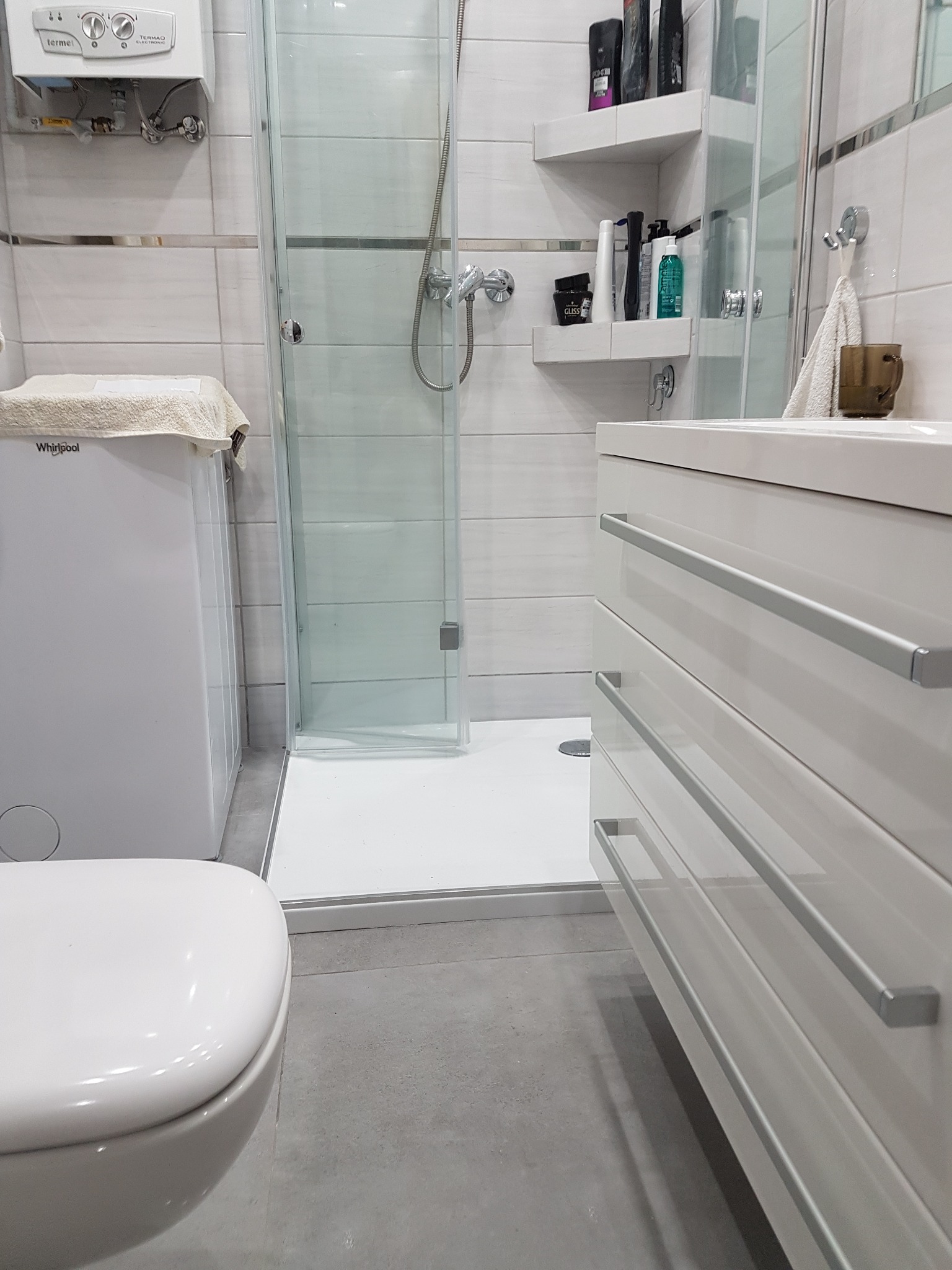 Nowoczesna łazienka z kabiną prysznicową typu walk-in, białą umywalką z szufladami z metalowymi uchwytami, wiszącą toaletą, grzejnikiem gazowym i półkami na kosmetyki.