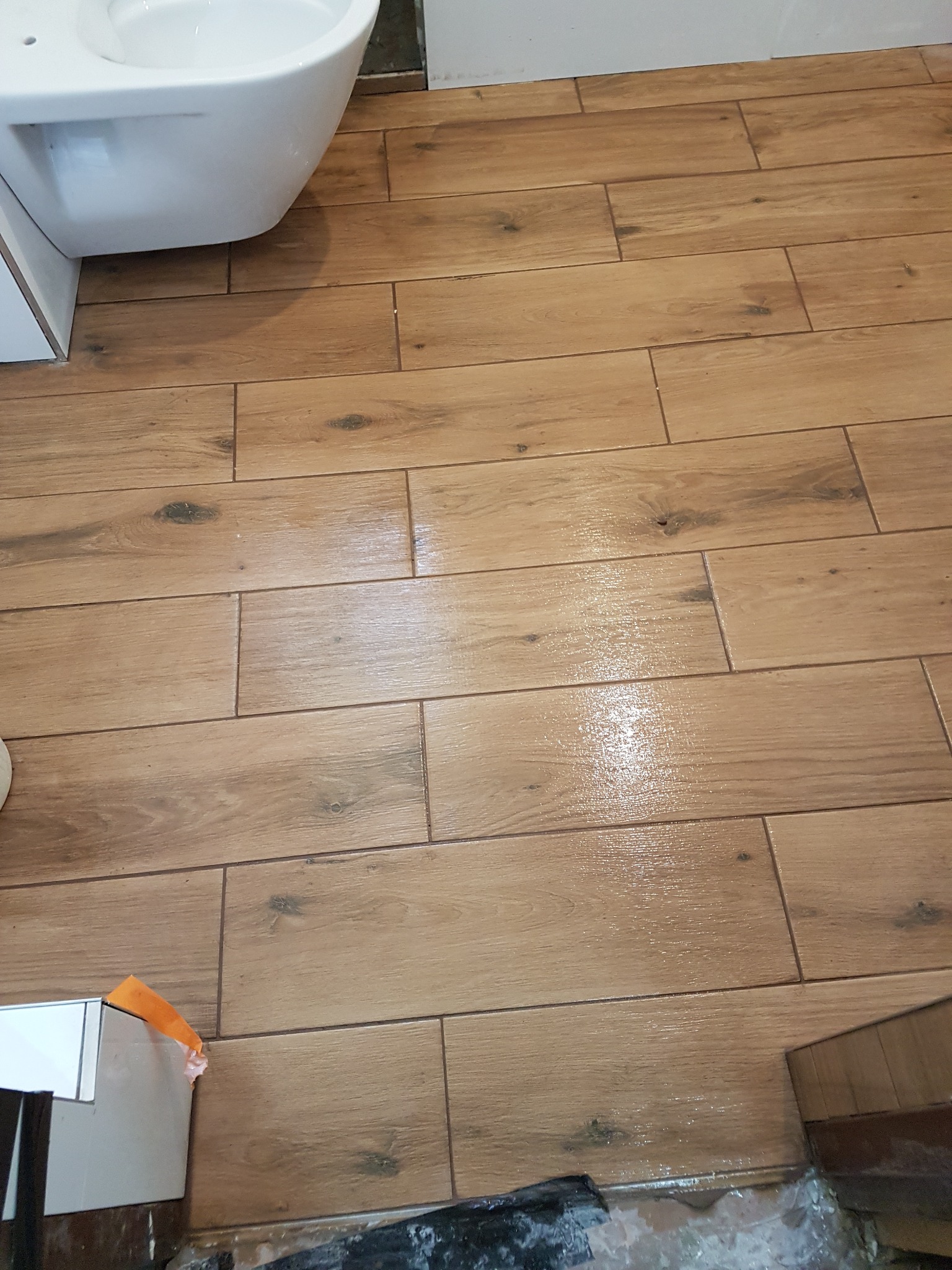 Ułożona podłoga z płytek imitujących drewno w łazience, widoczny fragment toalety i niedokończone wykończenie przy ścianie.