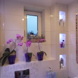 Jasna łazienka z białymi płytkami, trzema fioletowymi storczykami w doniczkach, wnękami z oświetleniem LED i lustrem w ozdobnej ramie.