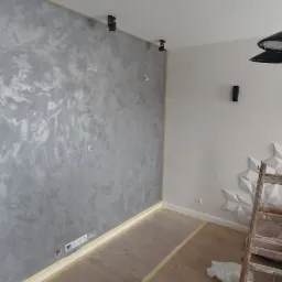 Ściana z efektem dekoracyjnym w odcieniach szarości, zabezpieczona folią podłoga, drabina oparta o ścianę z dekoracyjnymi panelami 3D, nowoczesna lampa sufitowa.