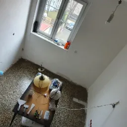 Pokój w trakcie prac przygotowawczych do malowania: ściany świeżo zagruntowane, na podłodze widoczne ślady farby, stół roboczy z narzędziami malarskimi i szpachelkami pod lampą.