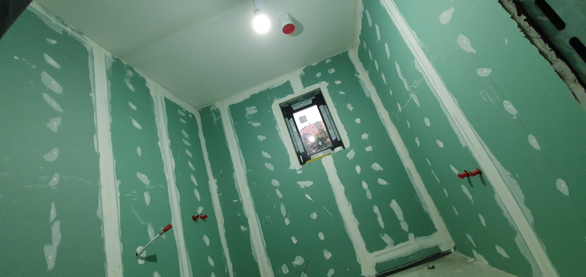 Wnętrze w trakcie prac: płyty karton gips na ścianach, widoczne łączenia zaszpachlowane, instalacja elektryczna wystająca ze ściany, okno z widokiem na zewnątrz.