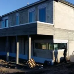 Surowy, dwukondygnacyjny dom w trakcie budowy, z widocznymi oknami z częściowo opuszczonymi roletami zewnętrznymi w kolorze białym, na tle błękitnego nieba.