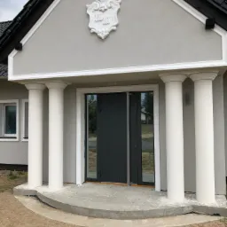 Wejście do domu z szarą elewacją, czarnymi oknami i drzwiami, wsparte na białych kolumnach, zwieńczone dekoracyjnym herbem.