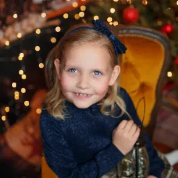 Uśmiechnięta dziewczynka z niebieskimi oczami, siedząca na żółtym fotelu, trzymająca lampion ze śnieżną kulą na tle choinki z lampkami.