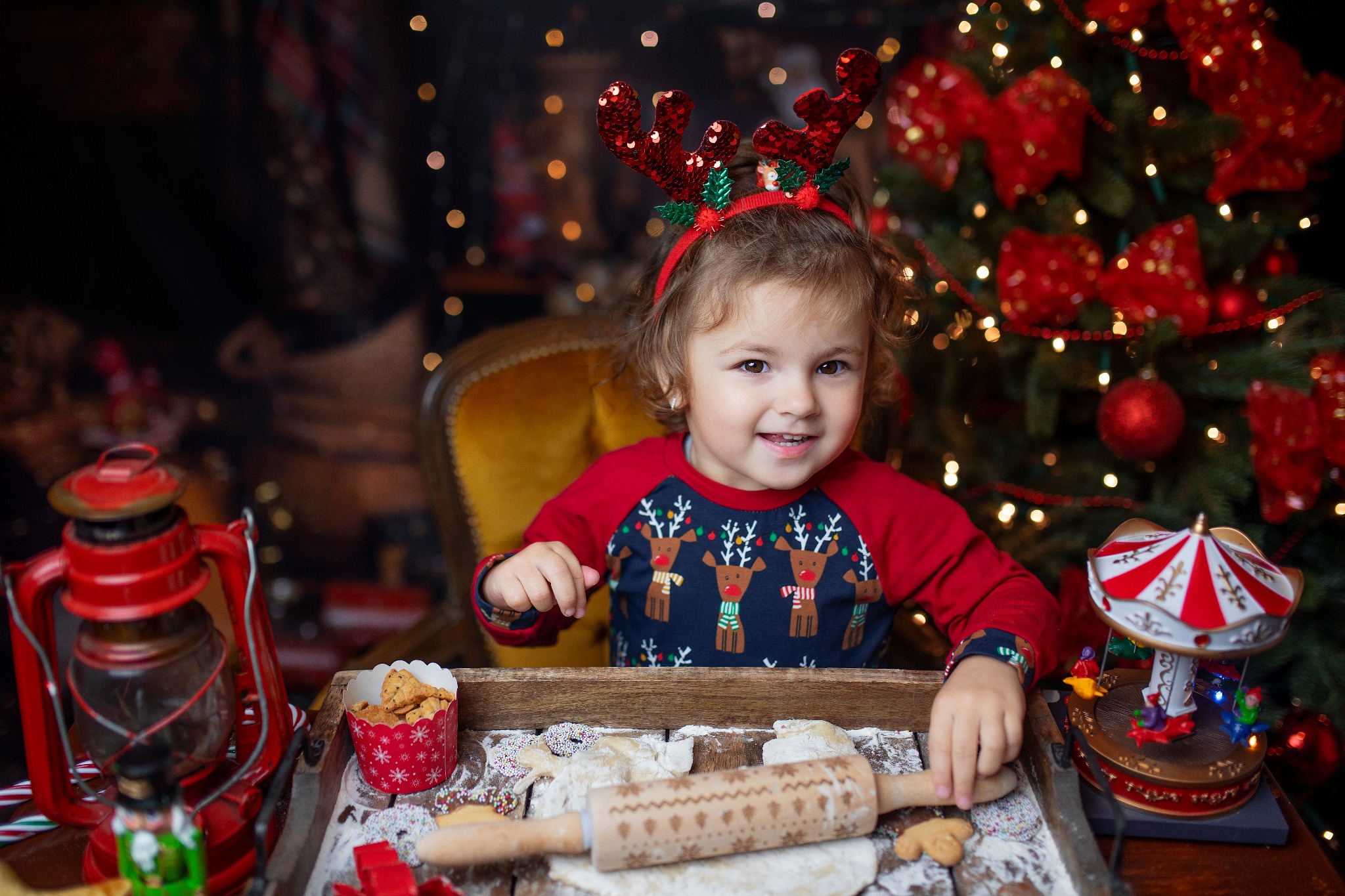 Uśmiechnięta dziewczynka w opasce z rogami renifera wałkuje ciasto na pierniczki na drewnianej tacy z dekoracjami świątecznymi, w tle choinka z czerwonymi ozdobami i lampkami.