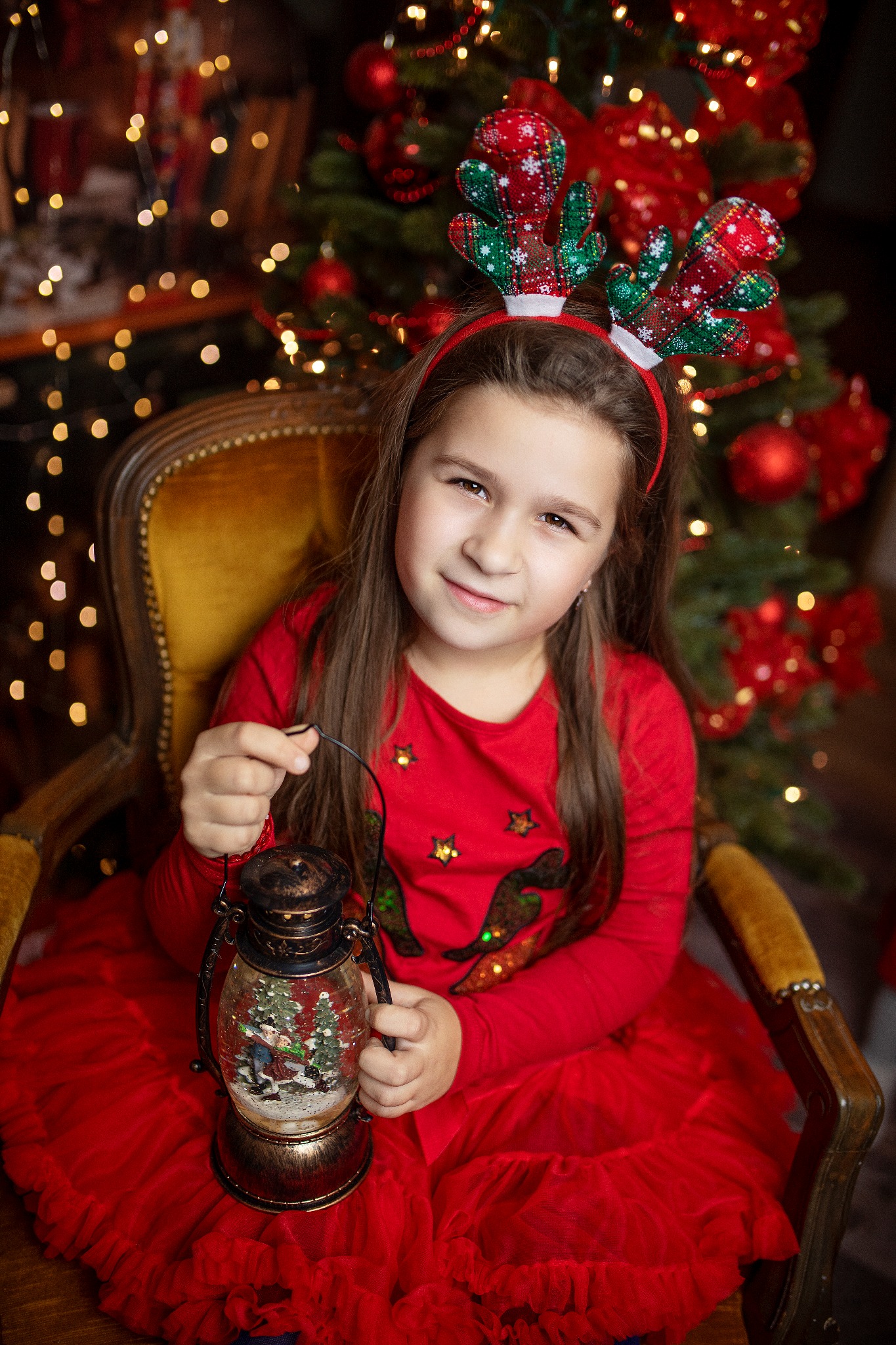 Uśmiechnięta dziewczynka w czerwonej sukience i opasce z rogami renifera siedzi na żółtym fotelu, trzymając lampion ze śnieżną scenką, na tle choinki z czerwonymi ozdobami i światełkami.