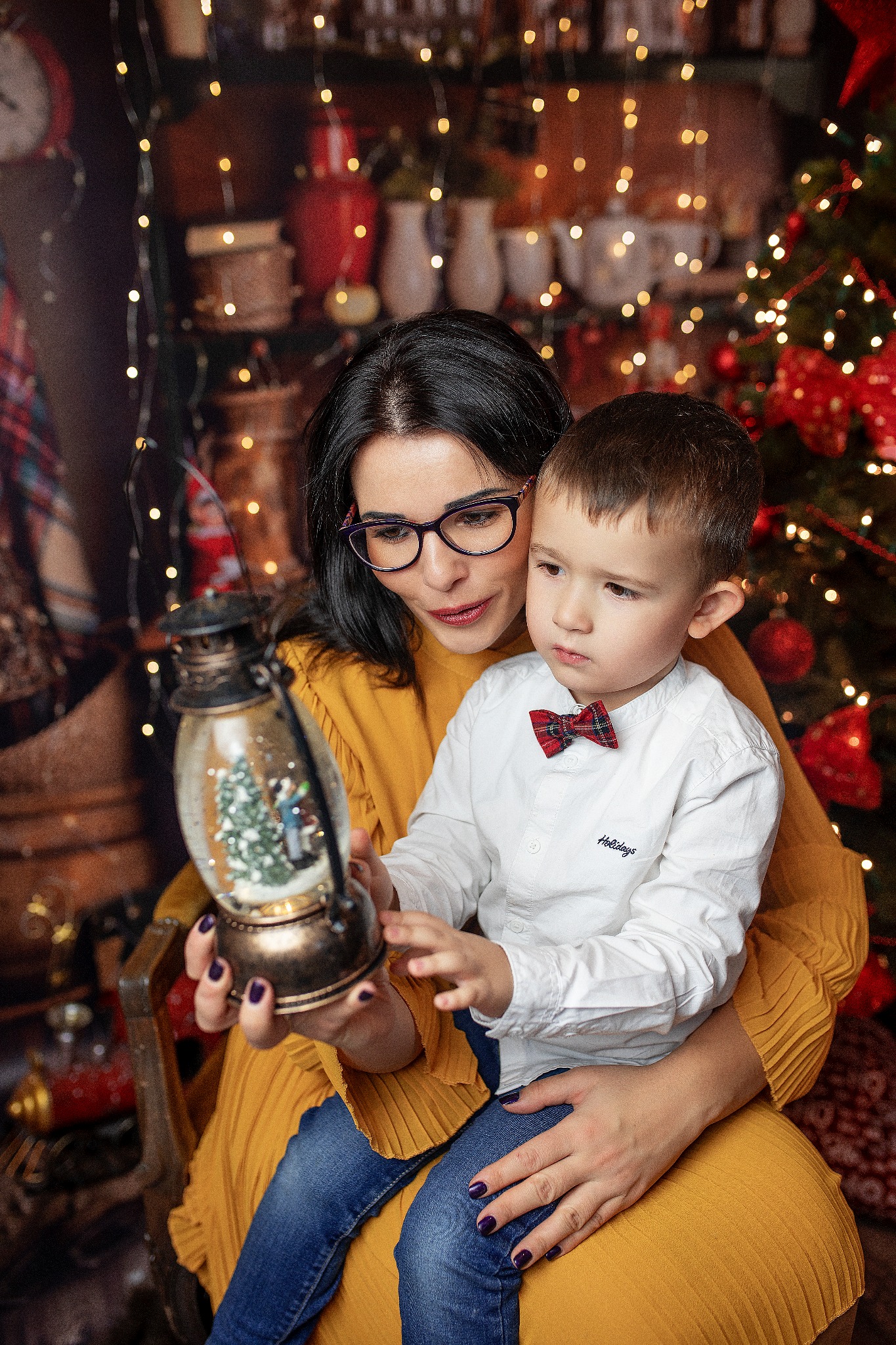 Kobieta w okularach trzymająca synka na kolanach, oboje oglądają szklaną kulę z zimowym krajobrazem, w tle choinka z lampkami.