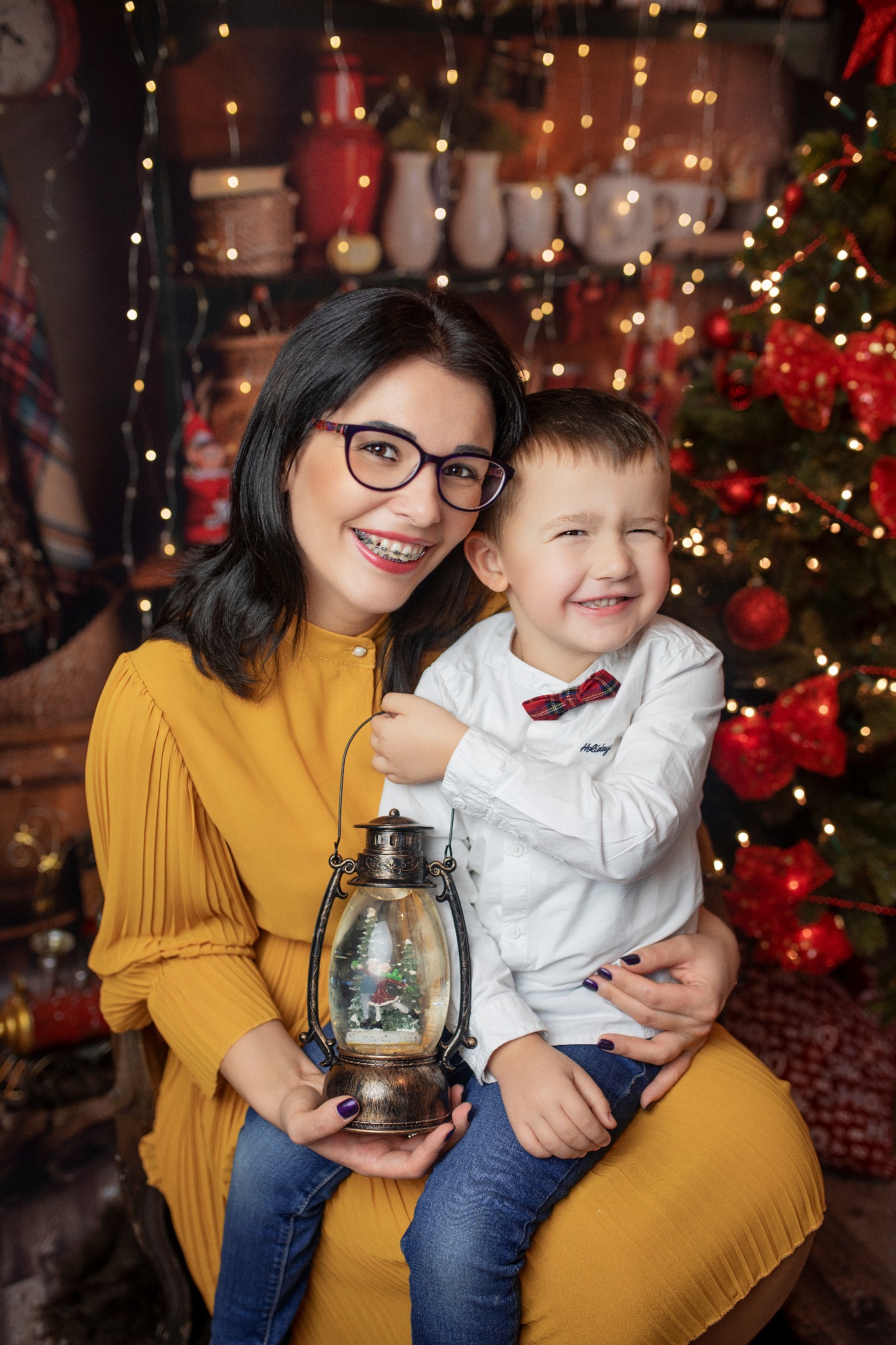 Uśmiechnięta kobieta w musztardowej sukience i okularach, z aparatem ortodontycznym, trzymająca lampion ze śnieżną kulą, pozuje z synkiem w białej koszuli i czerwonej muszce na tle świątecznej...