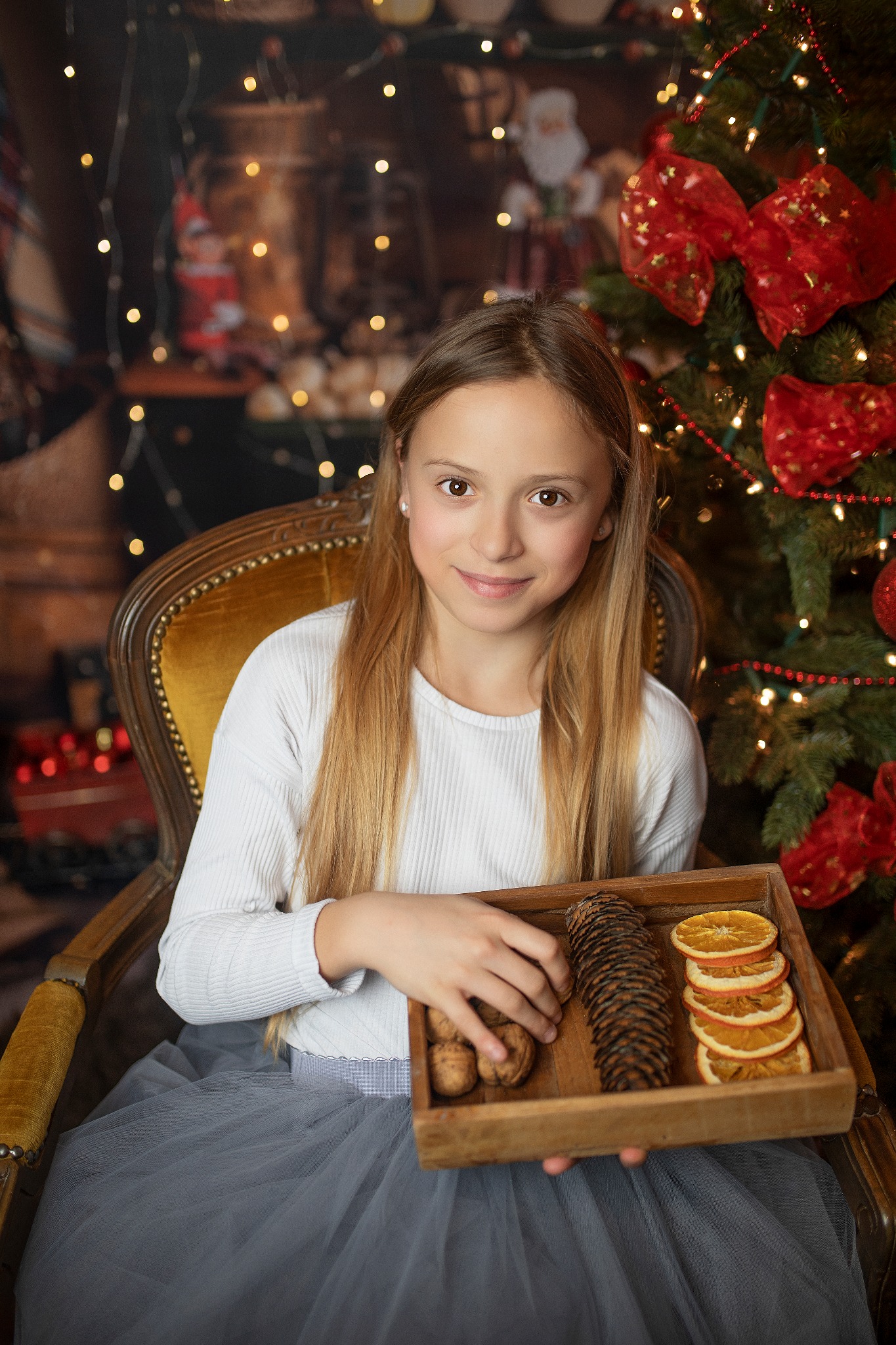 Dziewczynka w fotelu z drewnianą tacą pełną orzechów, szyszek i suszonych pomarańczy na tle choinki z czerwonymi kokardami i delikatnymi światełkami.