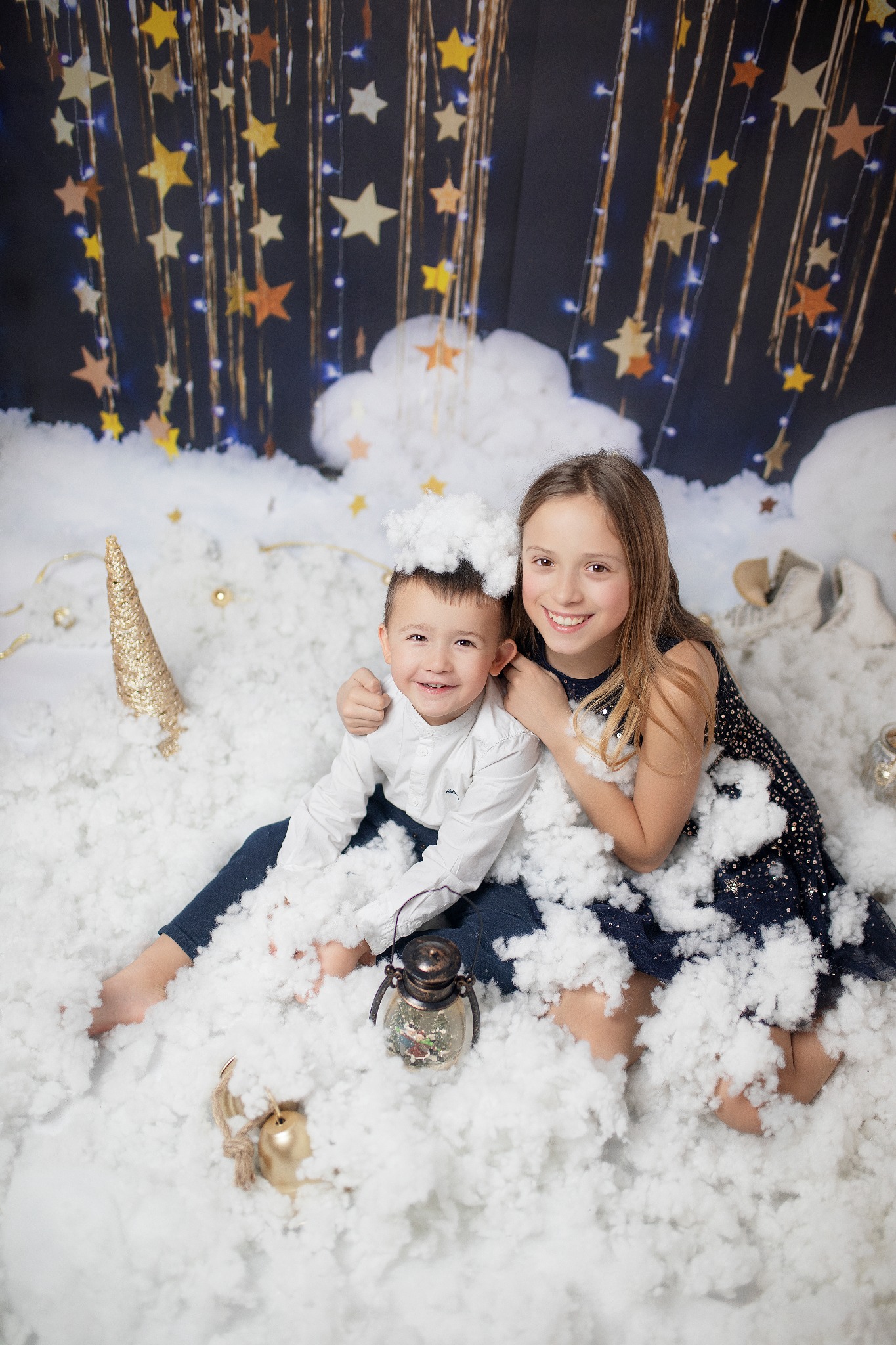 Uśmiechnięte rodzeństwo, chłopiec z watą na głowie, dziewczynka w granatowej sukience, siedzą w sztucznym śniegu na tle gwiazd i światełek, dekoracyjna lampka i złoty róg jednorożca obok.