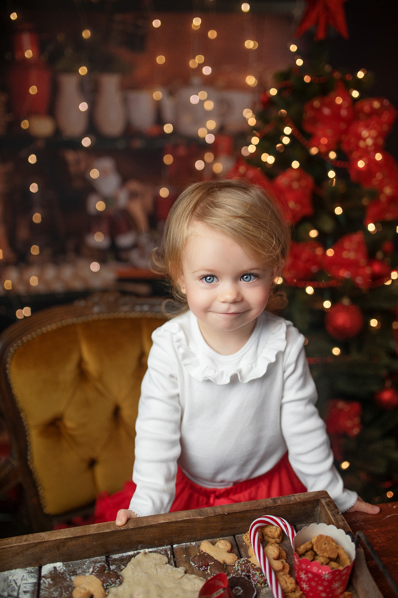 Uśmiechnięta dziewczynka z blond włosami i niebieskimi oczami w czerwonej spódniczce, opierająca się o drewnianą tacę z ciastkami i surowym ciastem, w tle choinka z czerwonymi ozdobami i światełkami.