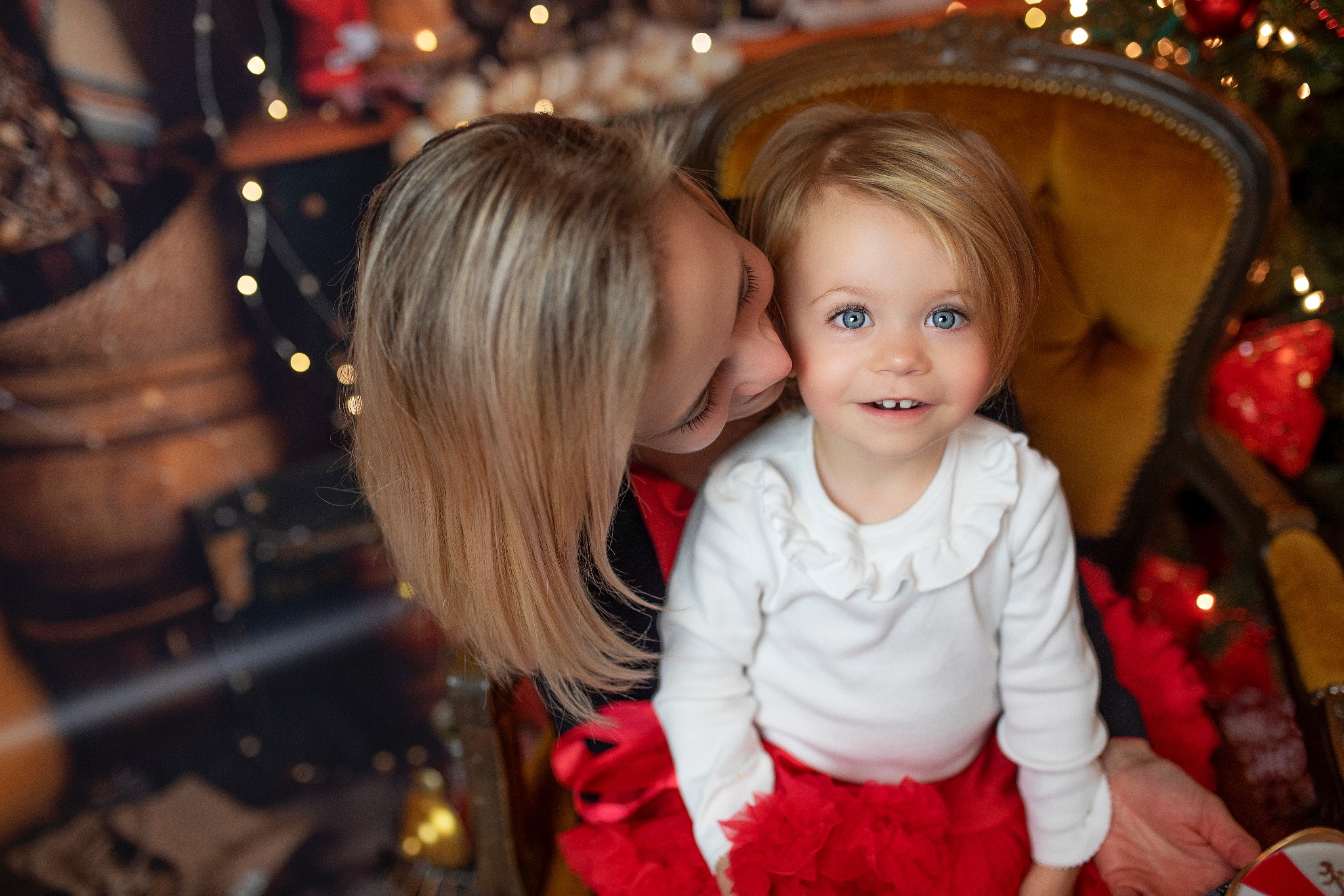 Blondwłosa matka całuje w policzek uśmiechniętą, jasnowłosą córeczkę o niebieskich oczach, siedzącą na żółtym fotelu na tle choinki z lampkami.